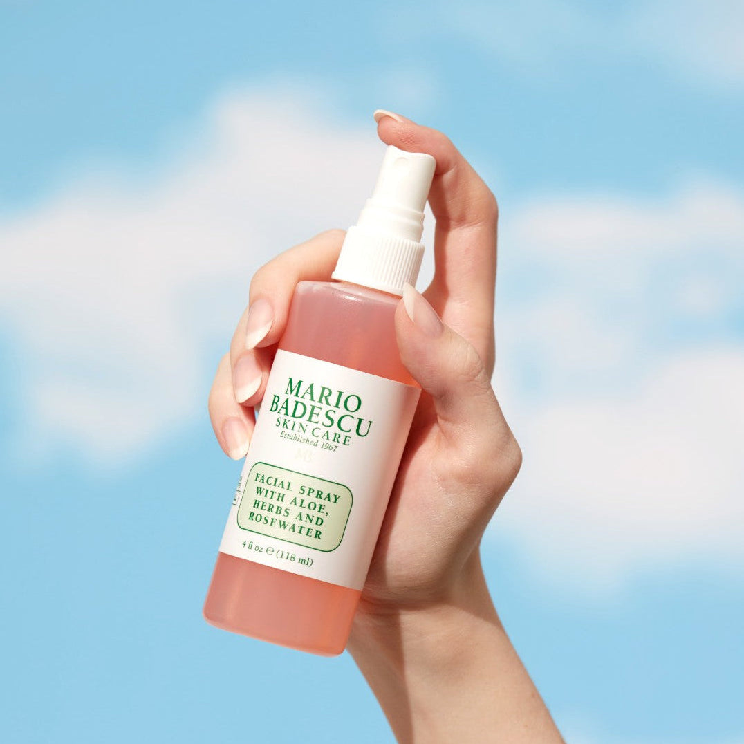 Hand holding a Mario Badescu facial spray against a blue sky background